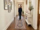 A woman walking barefoot on a blue and red patterned runner rug in a well-lit hallway with dark wood floors, white walls, gallery art, and a white console table with a mirror.