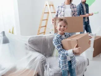 A young boy smiling while holding a cardboard moving box, with his parents carrying boxes and painting supplies in the background.