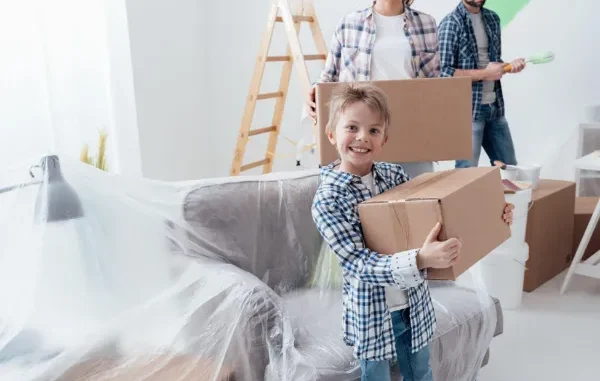 A young boy smiling while holding a cardboard moving box, with his parents carrying boxes and painting supplies in the background.